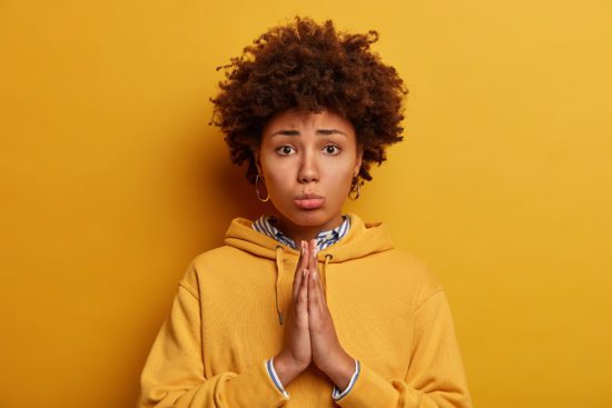 Let me please. Sad pleading African American woman asks for permission, holds hands in pray, says forgive me, poses against yellow background, wears sweatshirt. Begging and saying forgive me.